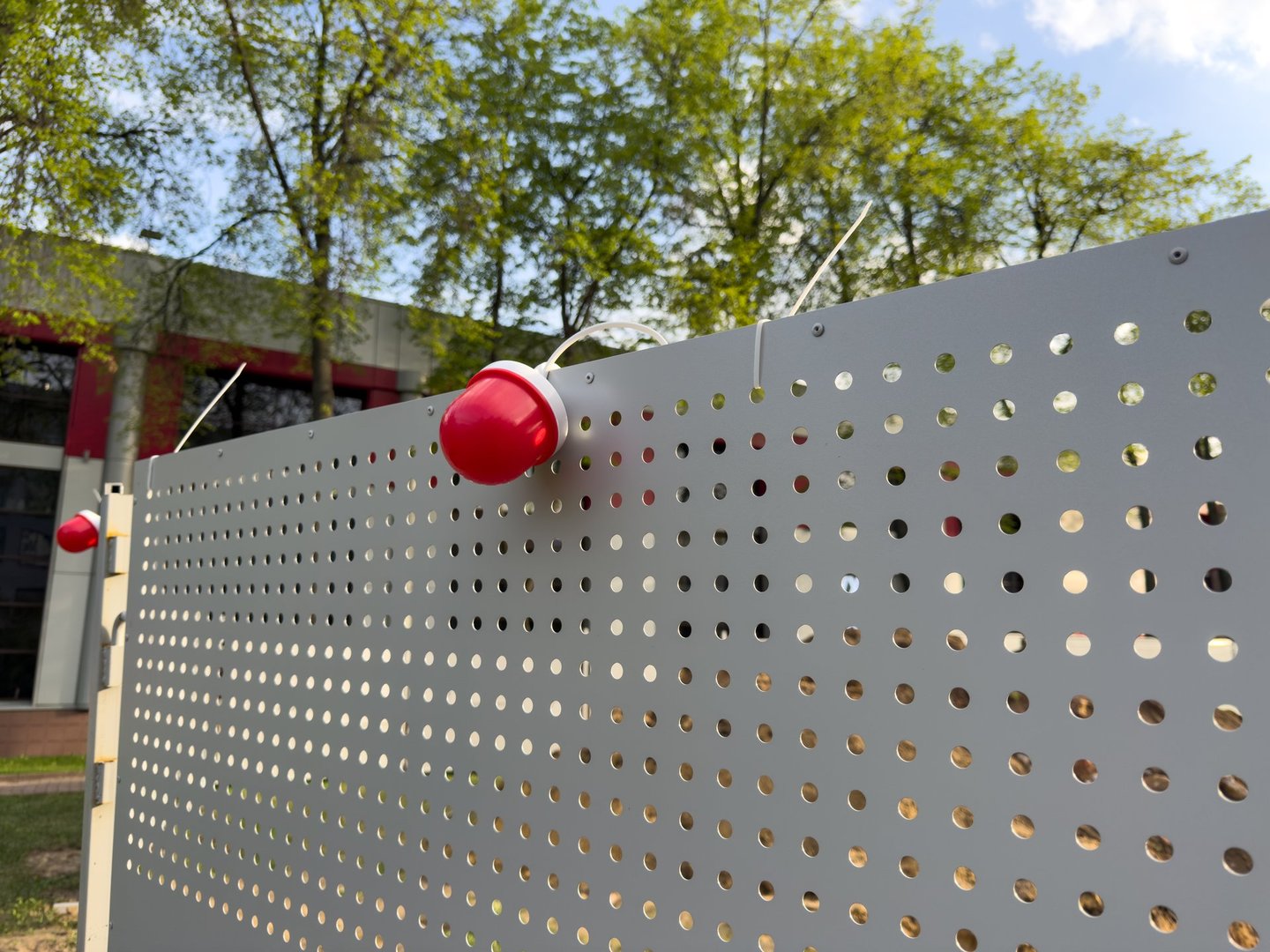 Modern perforated metal fence with red dome lights forms a sleek urban barrier near a contemporary public building. Functional design meets visual simplicity in this outdoor city infrastructure