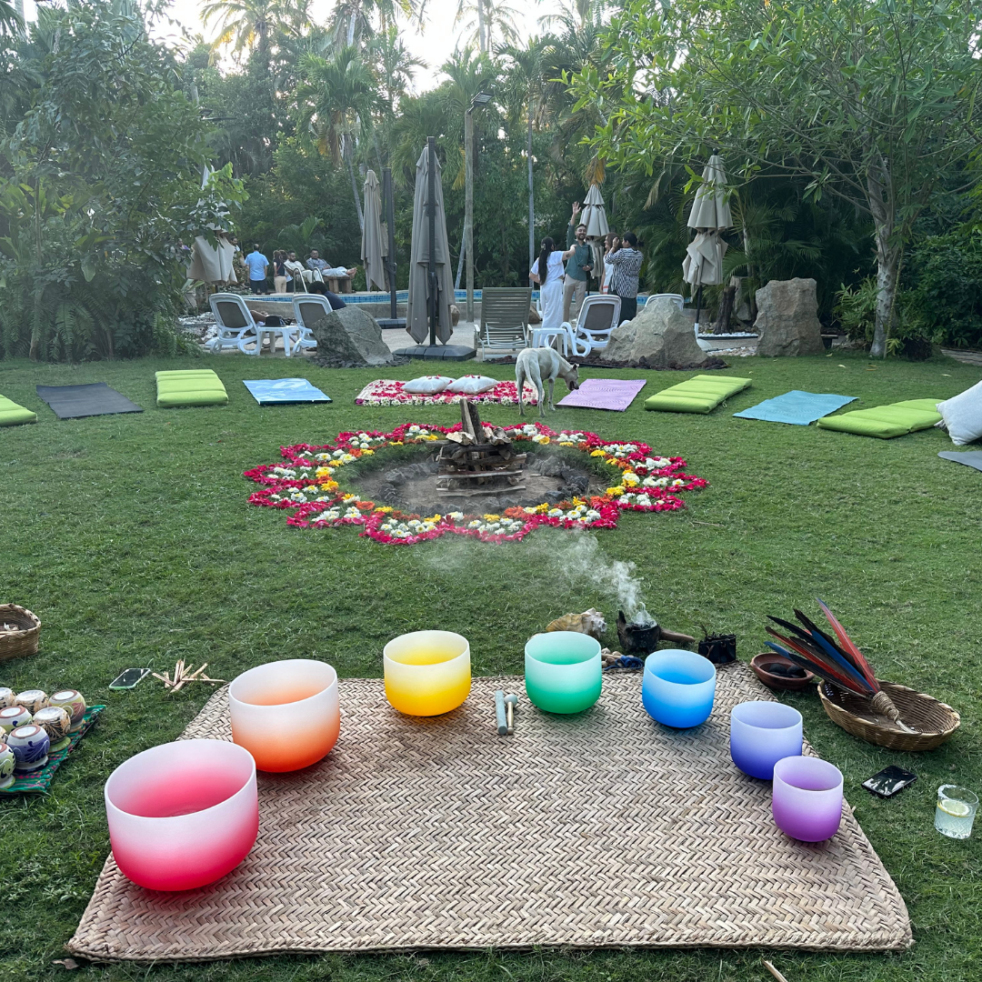 Outdoor meditation setup with colorful bowls, flower circle, cushions, and smoke rising, surrounded by lush greenery.