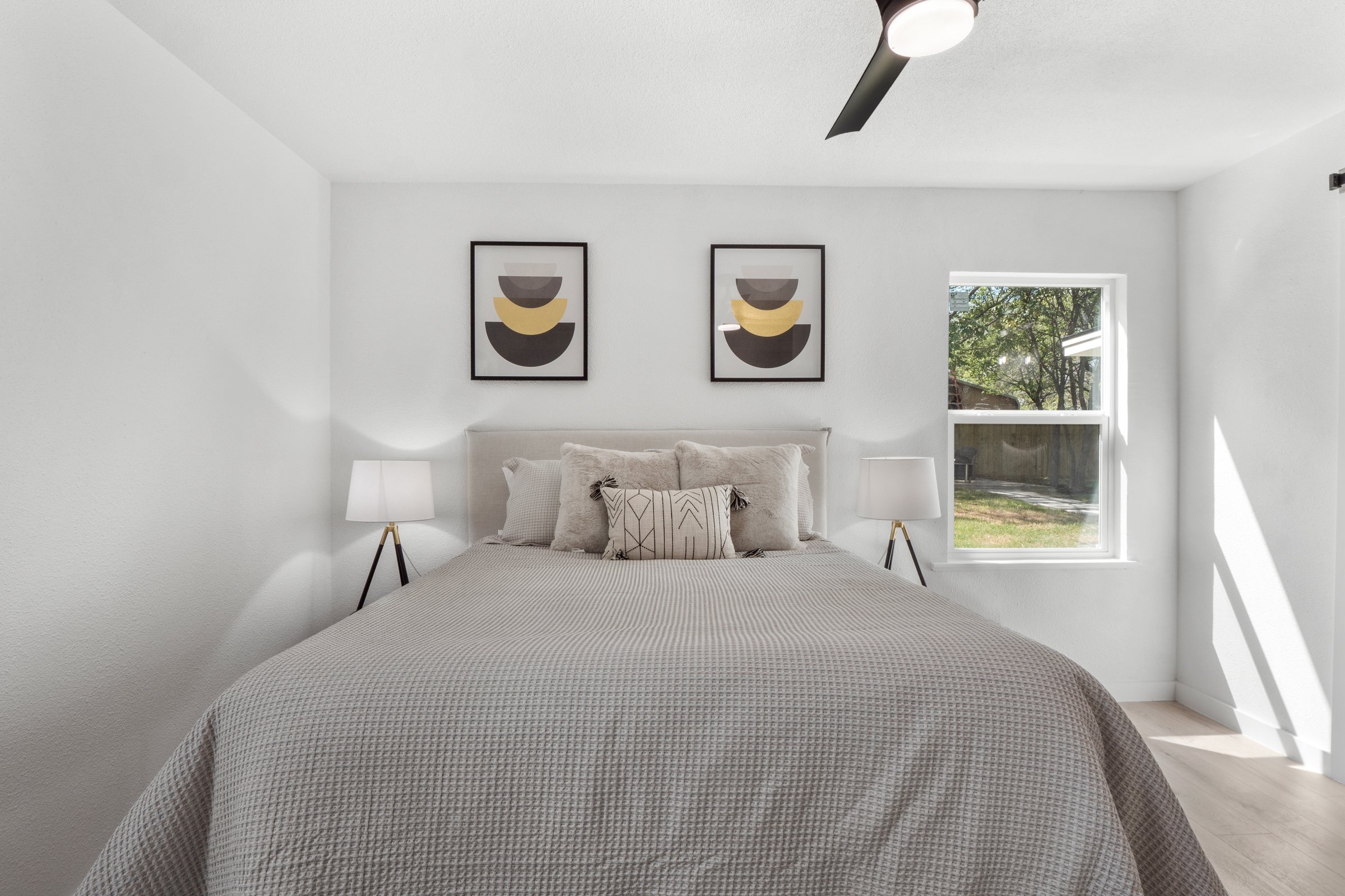 Minimalist bedroom with a neatly made bed, abstract art, and two bedside lamps, featuring a window with a backyard view.