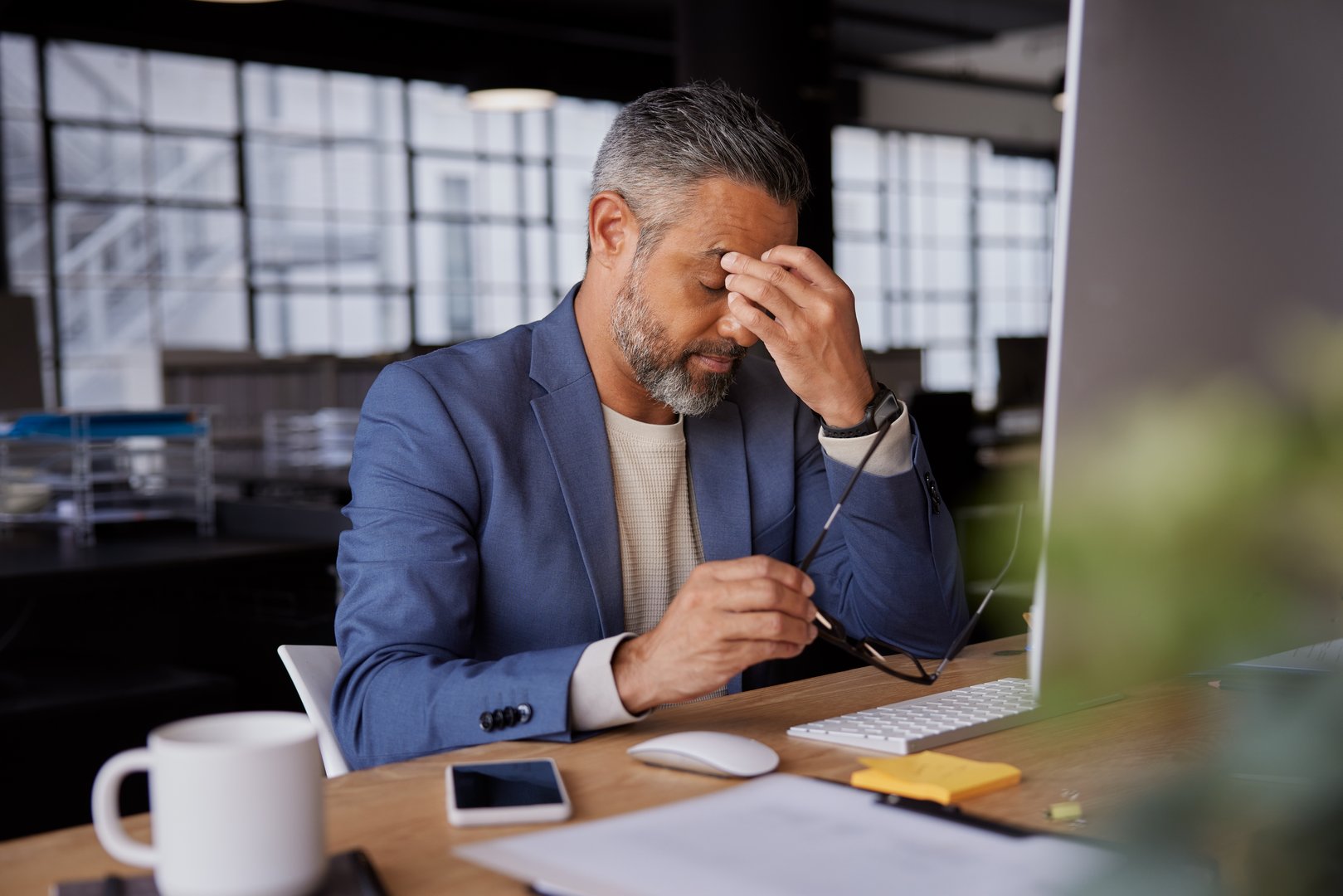 Mature businessman holding his head in stress, sitting at a desk with computer and documents. Indian manager working late and worried for company deadline. Stress, anxiety and burnout of a mid adult business man accounting risk and audit report or debt.