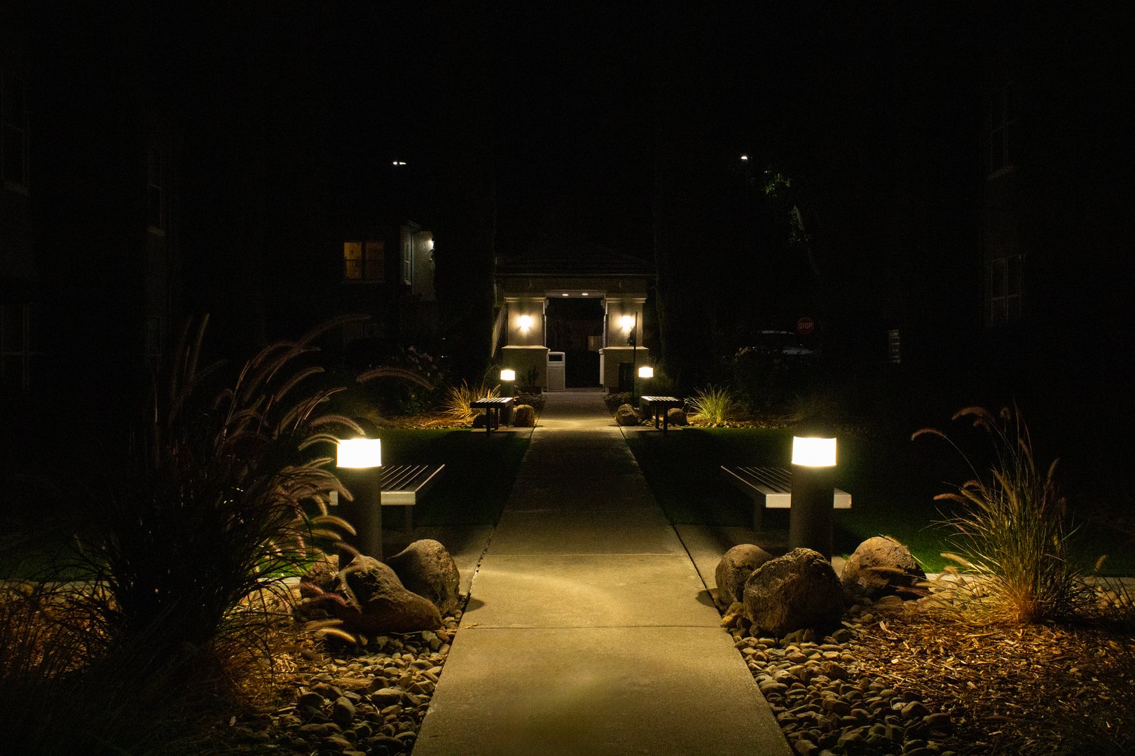 A peaceful pathway leads to a doorway, lit by soft lights on either side, surrounded by lush greenery and stones. Perfect for evening strolls or relaxation.
