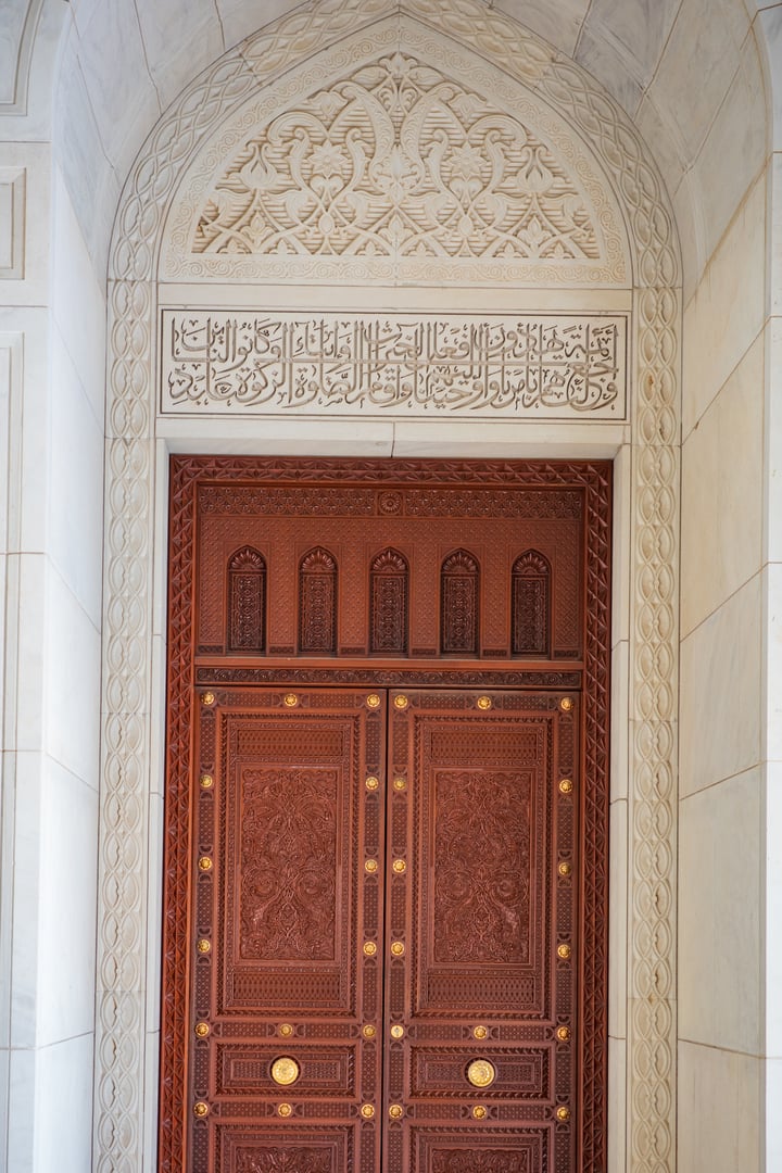 A detailed architectural photograph featuring an intricately carved wooden door framed by a beautifully decorated marble archway at the Sultan Qaboos Grand Mosque in Muscat, Oman. The composition showcases fine Islamic craftsmanship, including elegant Arabic calligraphy, floral patterns, geometric borders, and ornate woodwork with gold accents.