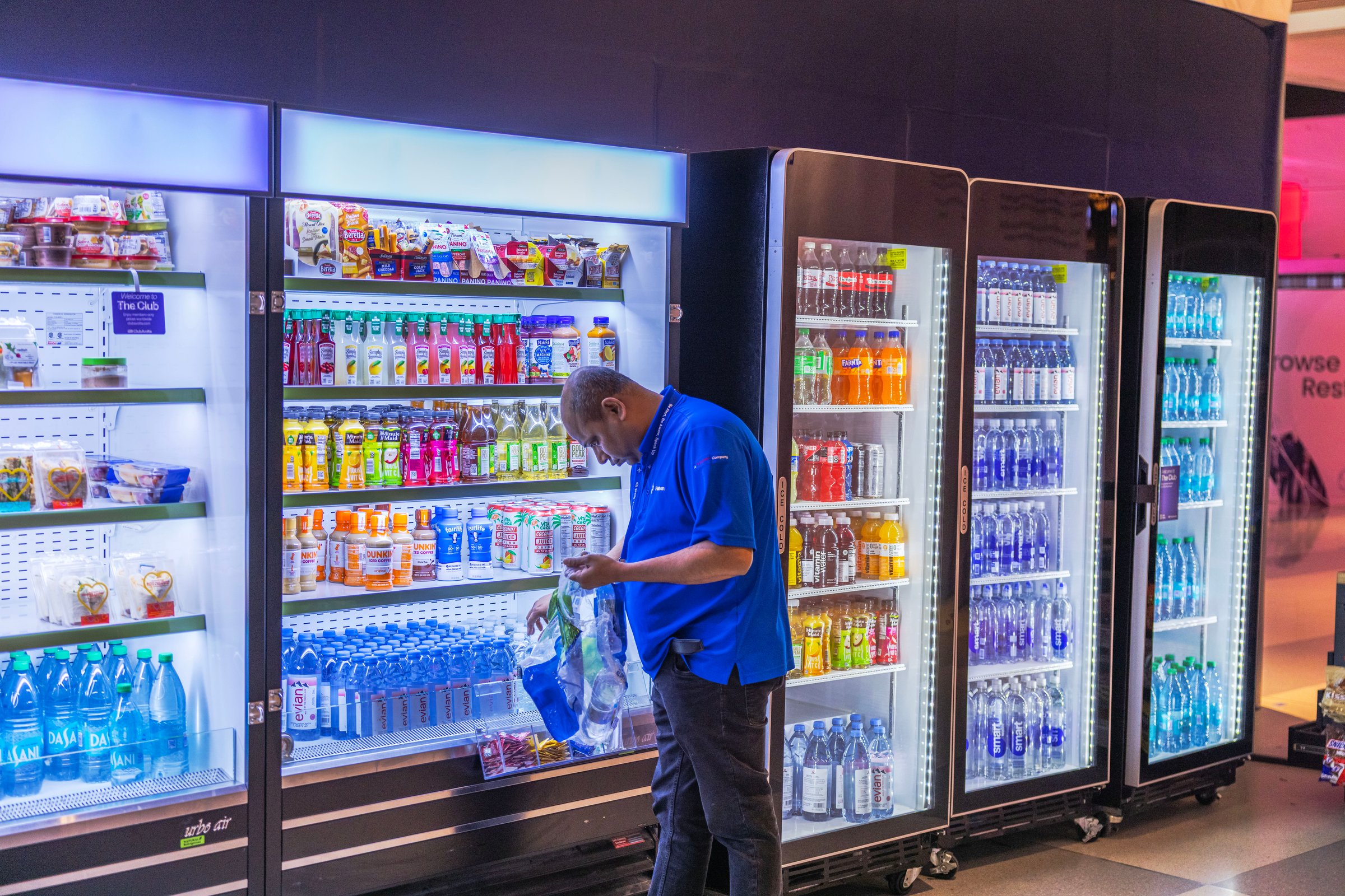 New York. USA. 04.06.2025. Worker restocking drink shelves in JFK airport shop with refrigerators full of beverages and snacks. .