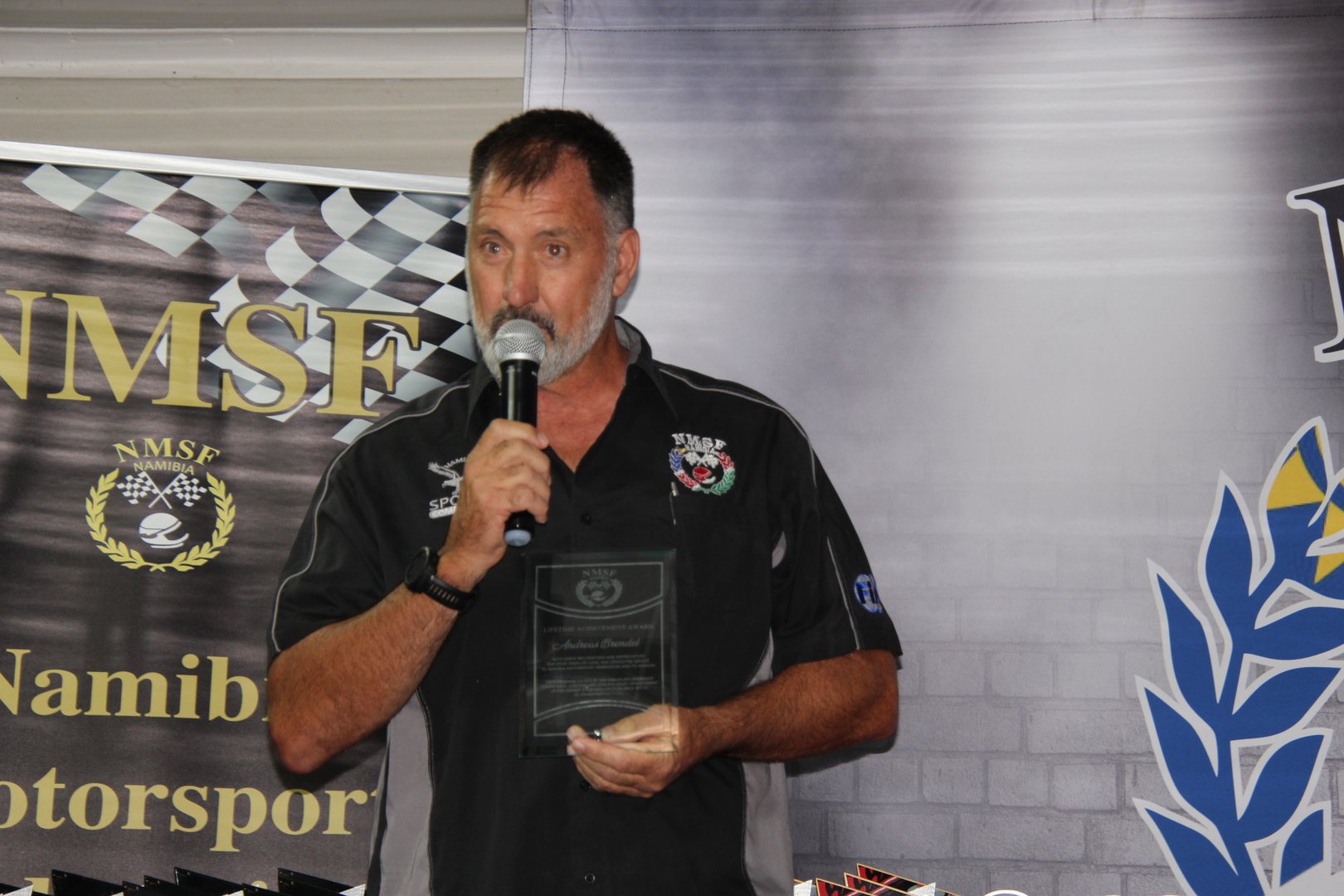 Man holding a plaque while speaking into a microphone at a motorsport event.