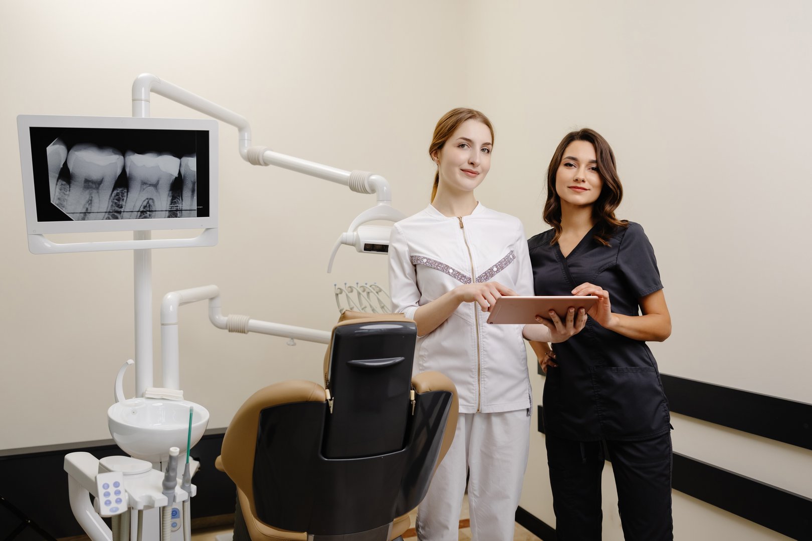 Two women dentist doctors are standing in a dental office and looking at a tablet.