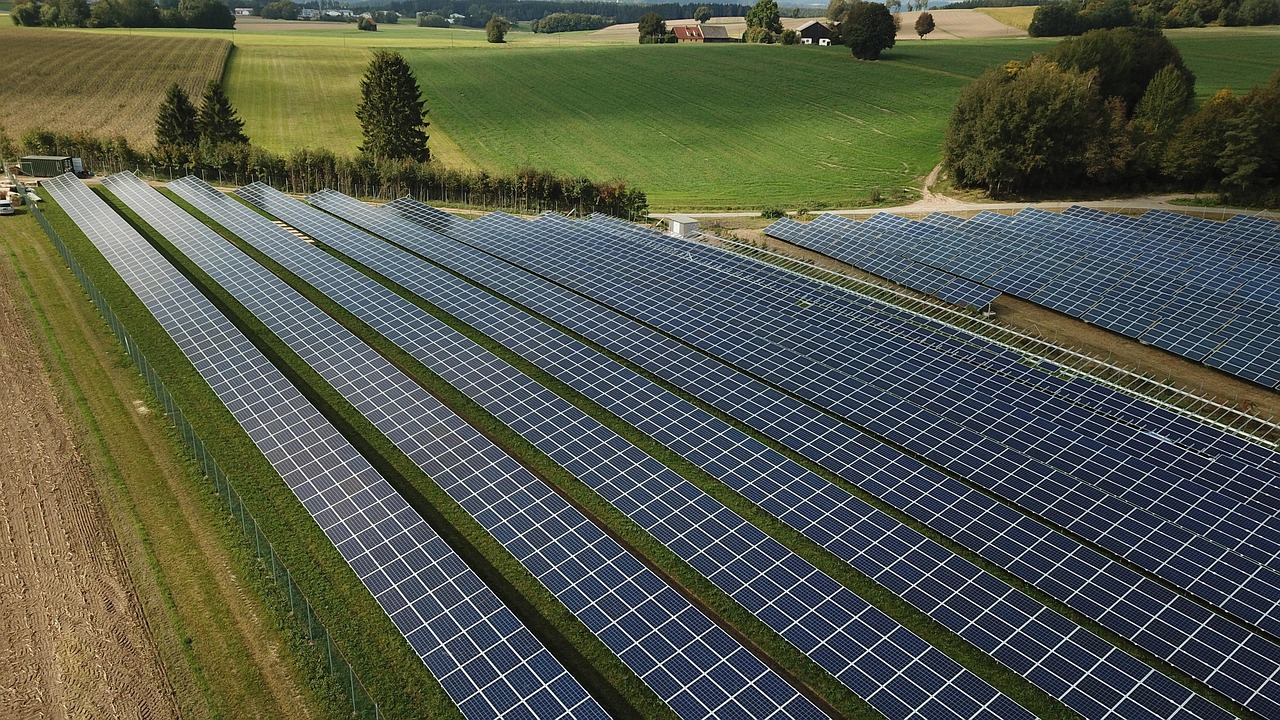 A large solar farm with rows of solar panels in a rural landscape, surrounded by fields and trees under a clear sky.
