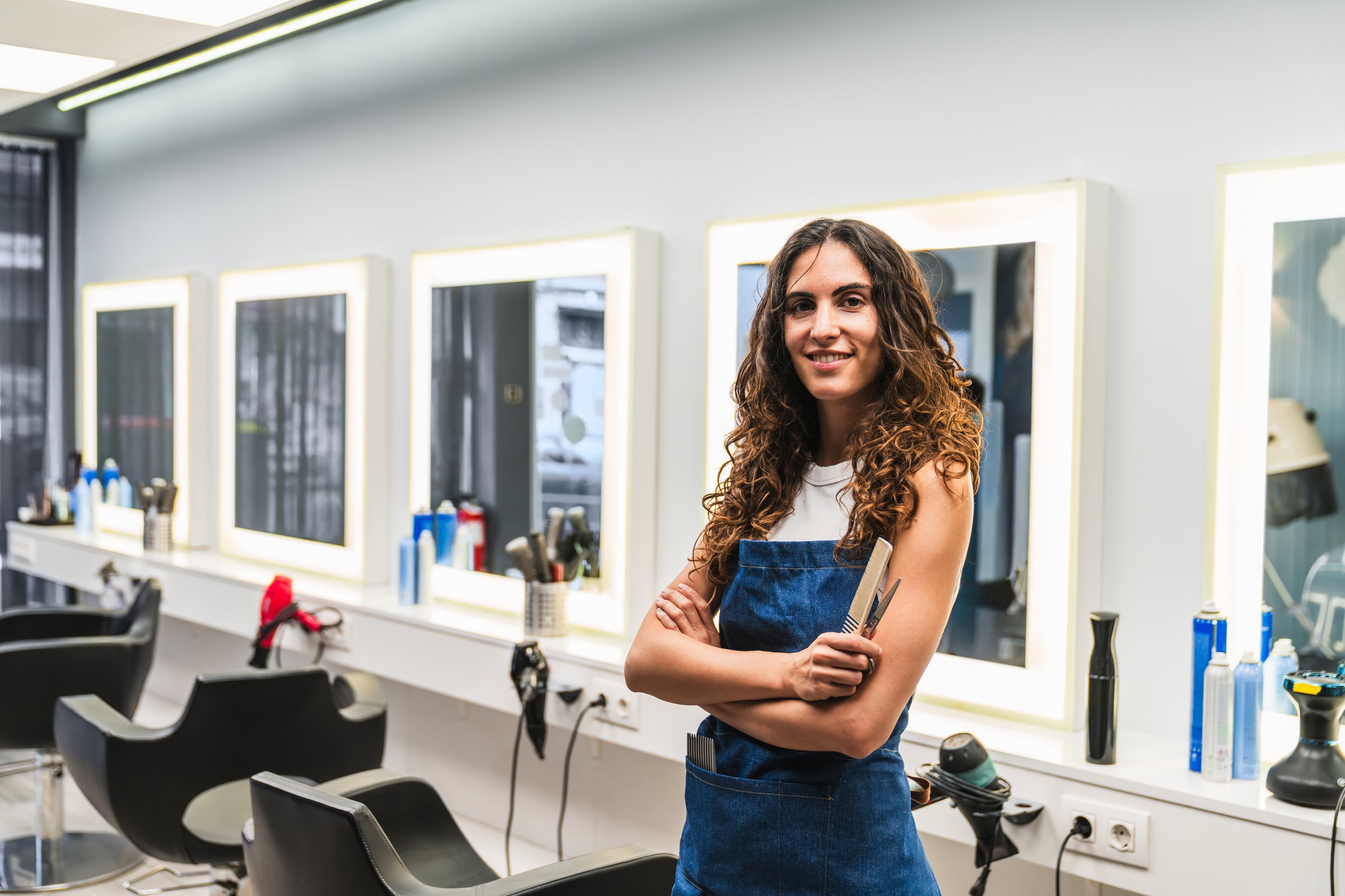 Smiling young woman, a professional hairstylist, standing confidently with arms crossed in her modern hairdressing salon