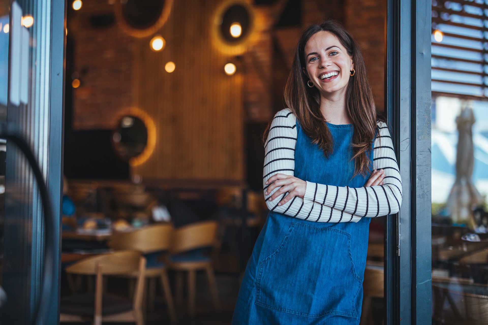 A joyful woman in a striped shirt and denim apron stands with arms crossed at a welcoming coffee house, embodying professional hospitality.