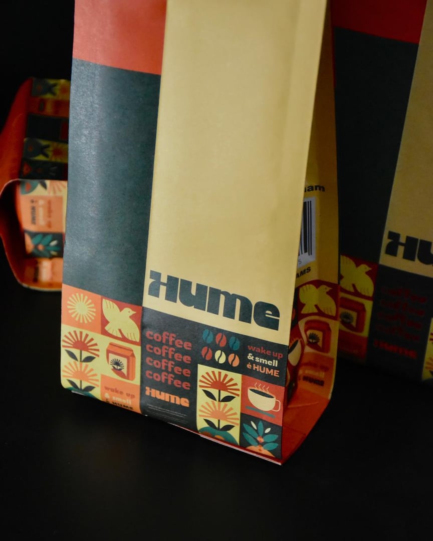 Colorful Hume coffee bags with floral and coffee cup designs, featuring the phrase "wake up & smell Hume" on a black background.