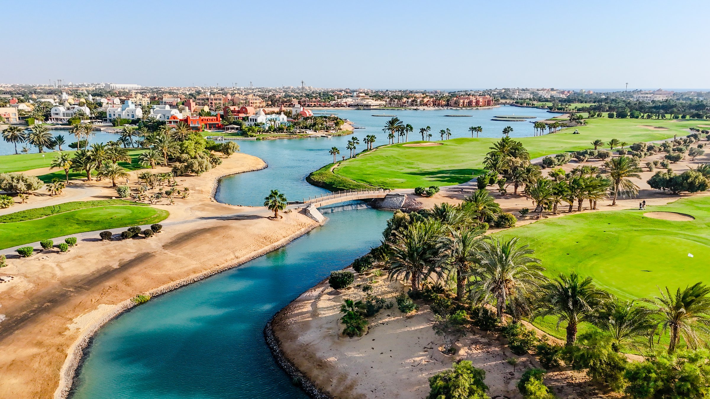 Expansive aerial view via drone overlooking the lush El Gouna golf course, clearly showing its integration with the surrounding resort town. Multiple fairways, palm trees, and blue lagoons are visible alongside residential villas and buildings under a clear sunny sky on the Red Sea coast of Egypt. Depicts the scale of the luxury tourism and residential destination.