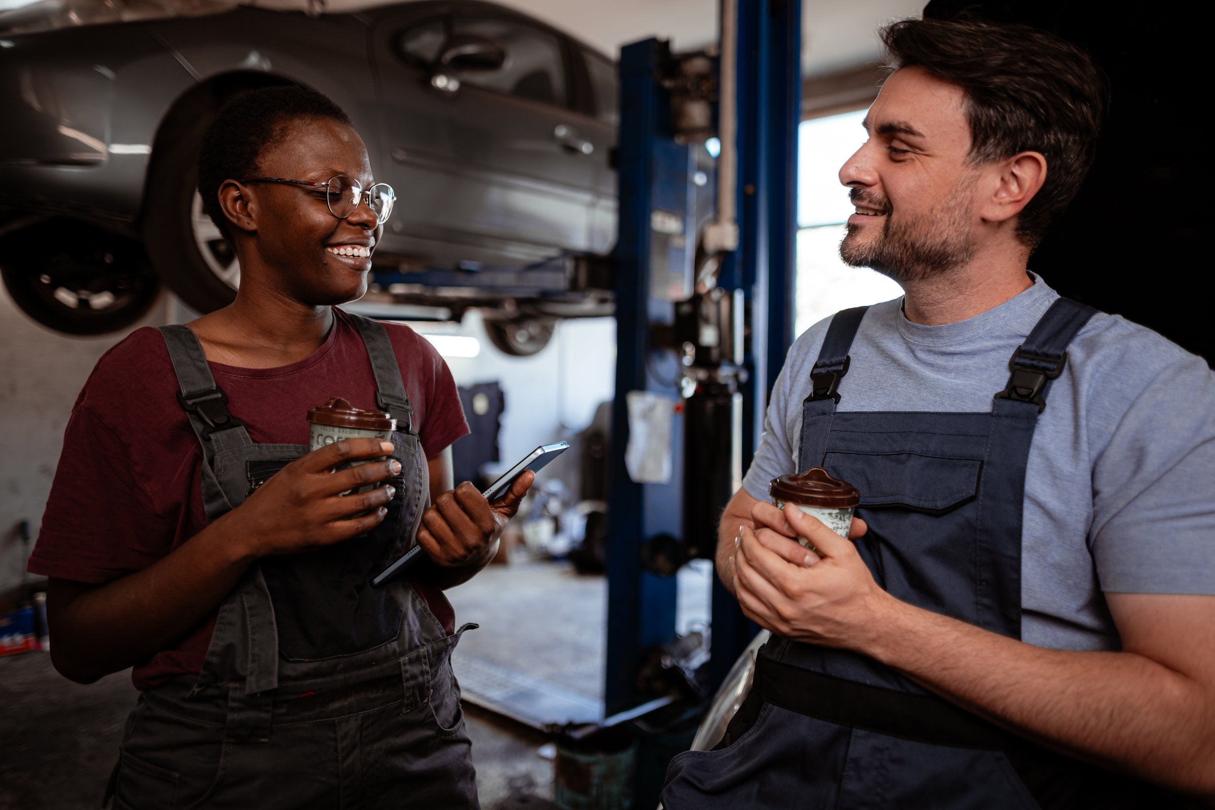 Two mechanics share a moment in a workshop, enjoying snacks and discussing car repairs with a vehicle elevated nearby.