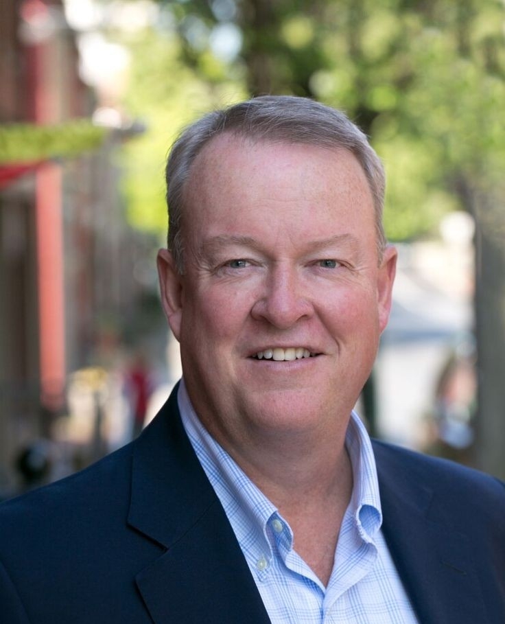 Smiling man in a business suit outdoors, standing on a leafy street, with blurred buildings in the background.