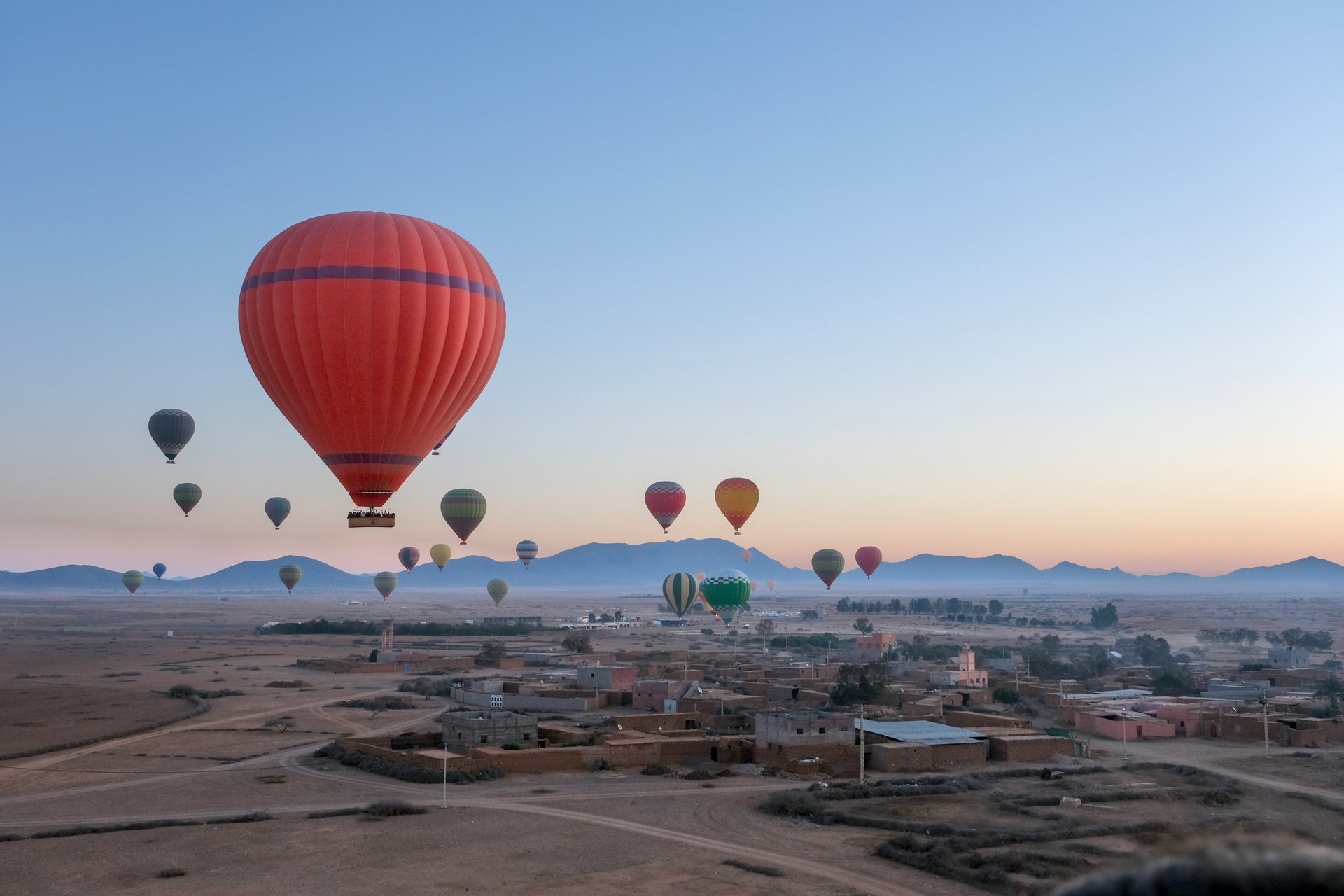 Hot air balloons float above an open landscape at sunrise, with mountains in the background. The sky transitions from deep blue to warm hues of dawn.