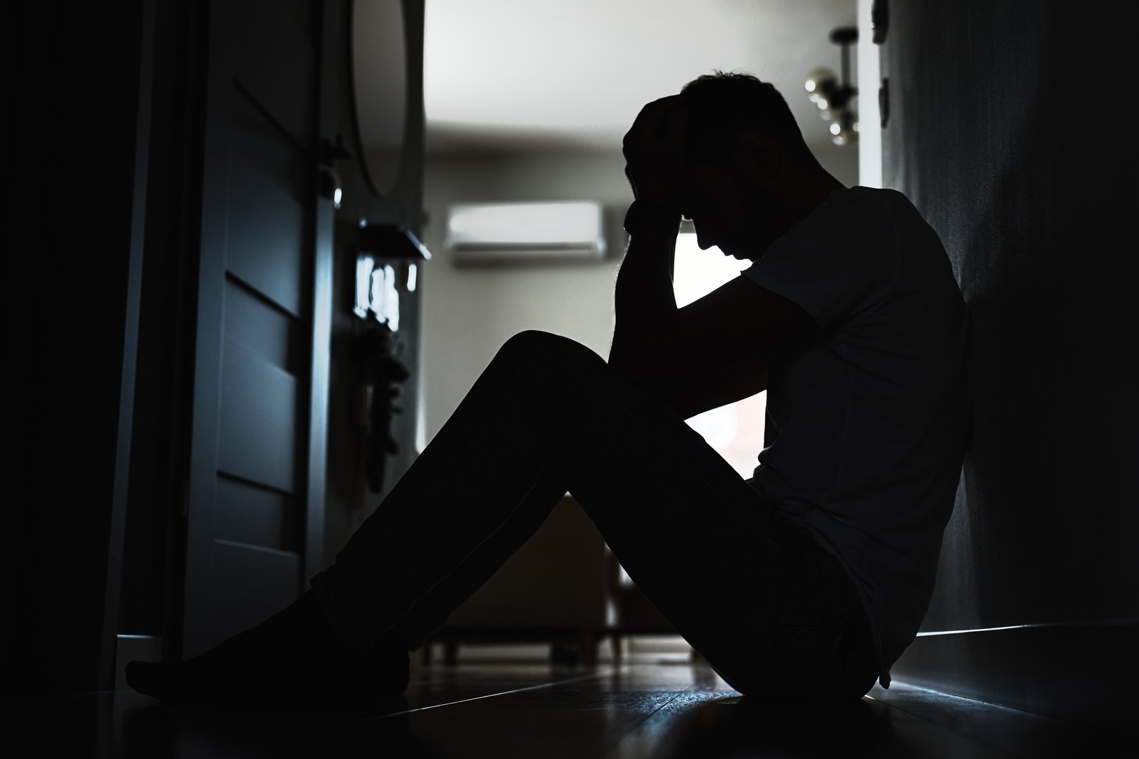 Depressed young man sitting on floor in dark room. Silhouette of young male suffering from loneliness and anxiety, covering his face with hands. Person with mental disorder