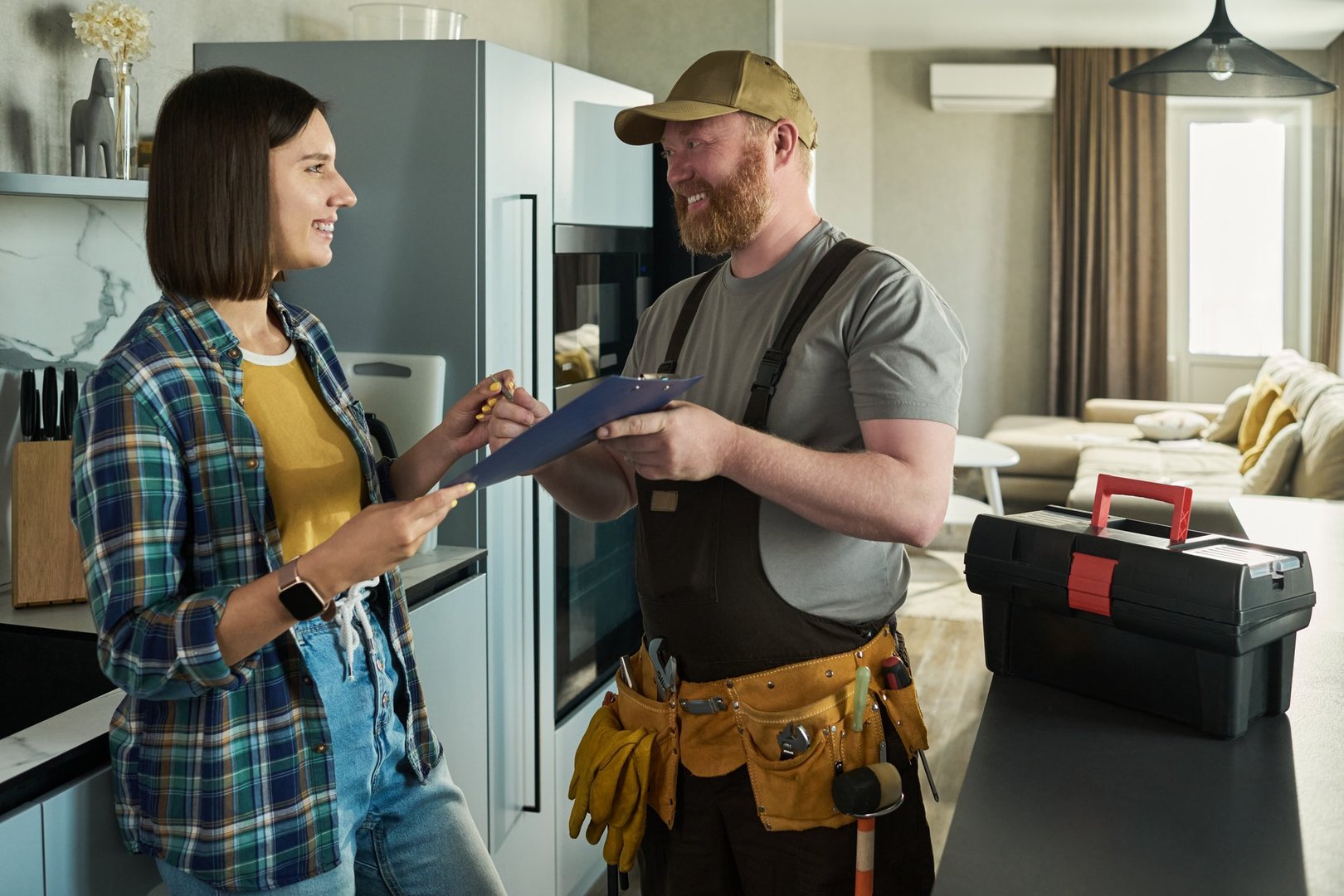 Caucasian young adult woman smiling while signing document held by Caucasian middle aged male repairman wearing tool belt, standing together in modern kitchen near toolbox