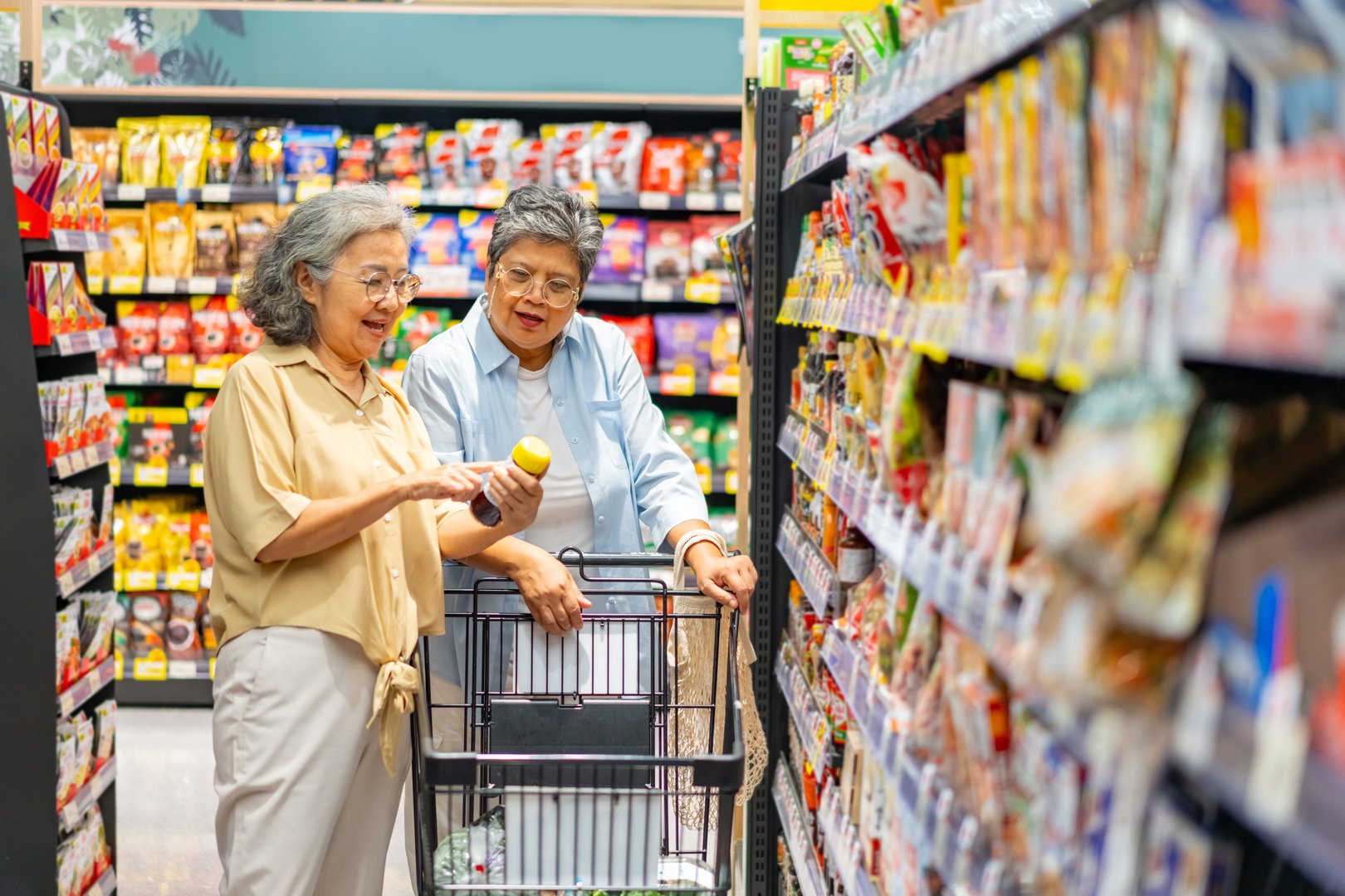 Happy Asian senior woman shopping at supermarket. Healthy elderly women friends enjoy urban lifestyle reading label, choosing and buying organic safety food, vegetable and groceries at shopping mall.
