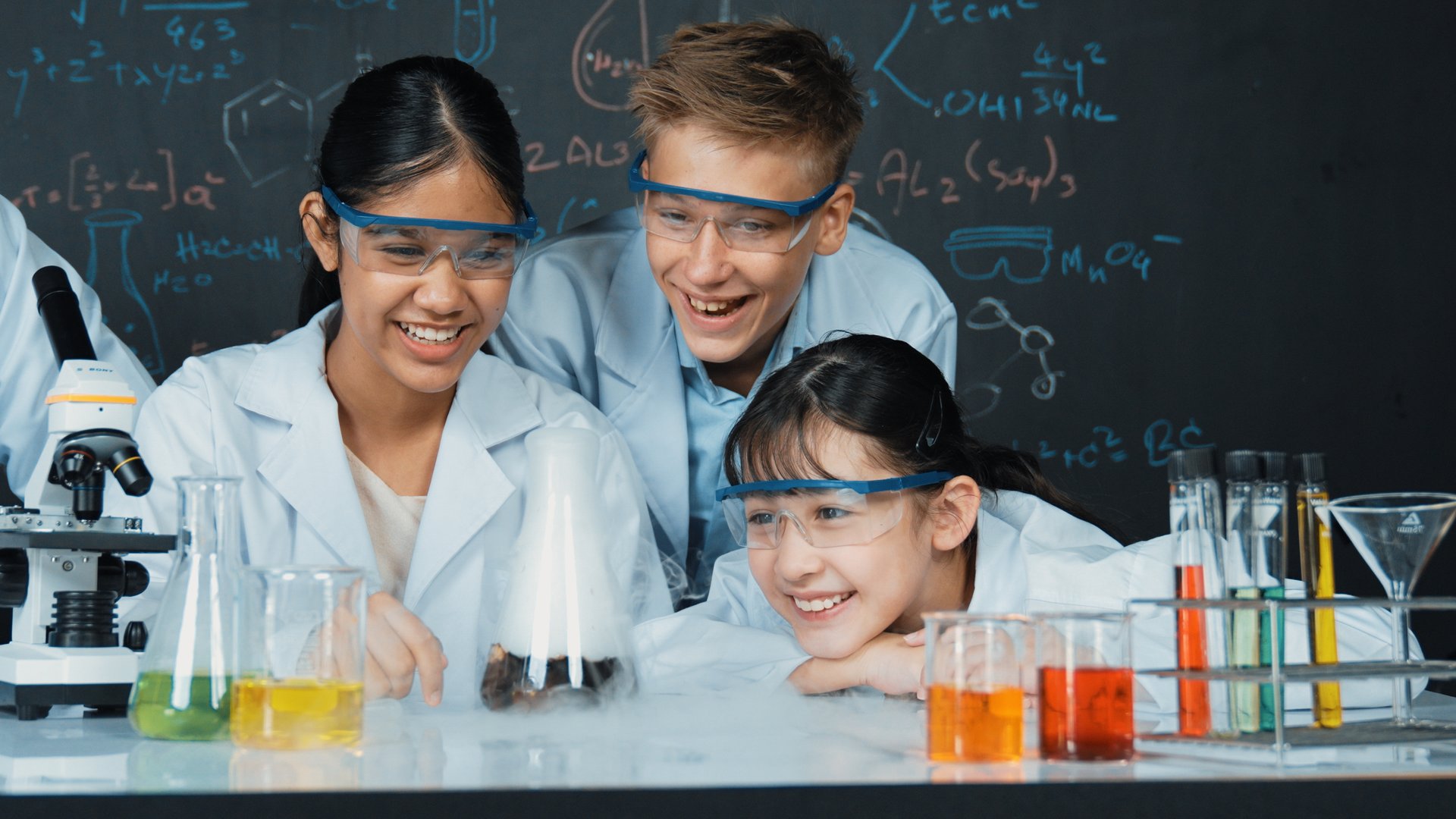 Girl looking at beaker while doing experiment at blackboard with chemistry theory in STEM class. teacher and group of highschool student with mixed races doing science activity together. Edification.