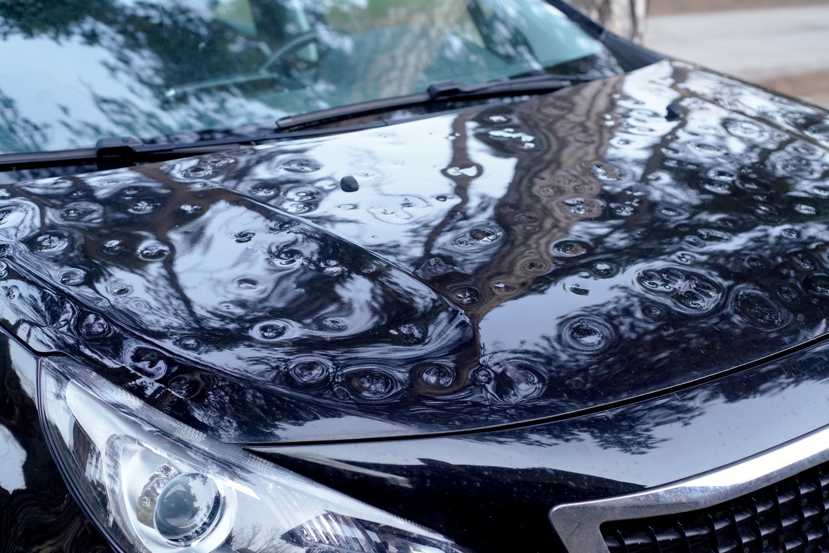 Close-up view of the hood, front pillar, and part of the roof of a car heavily damaged by hail. Highlights the importance of covered parking and auto insurance.
