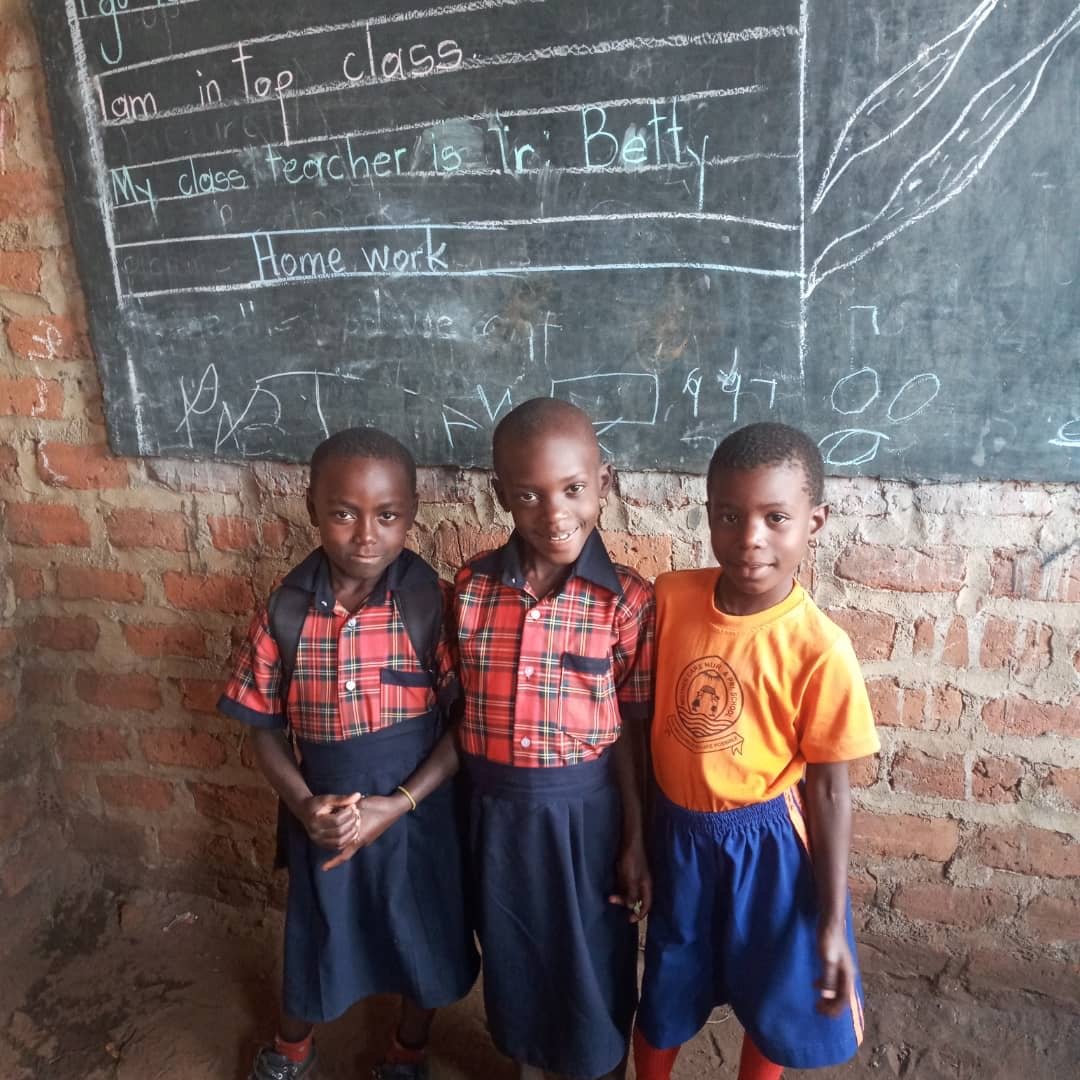 Three young children in school uniforms standing inside a classroom in front of a chalkboard with writing on it.