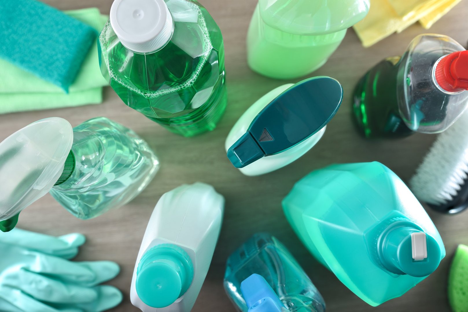 Supply of general cleaning products with several disinfectant bottles and tools such as rags, scouring pads, gloves and brushes on wooden table. Top view.