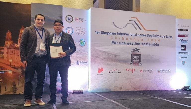 Two individuals standing in front of a backdrop at a symposium. One holds a certificate. Event banners are visible.