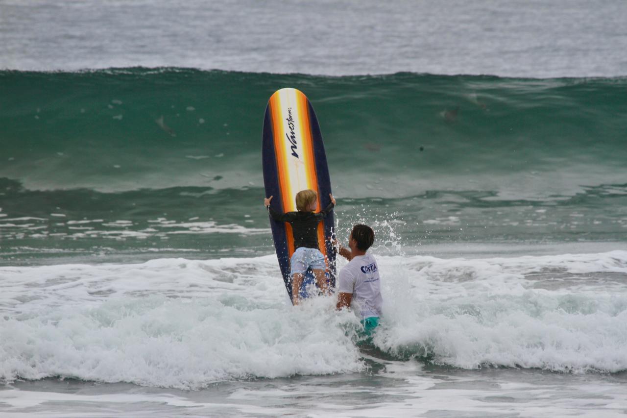 A child with a surfboard walks toward the ocean waves, accompanied by an adult in the water, both preparing to surf.
