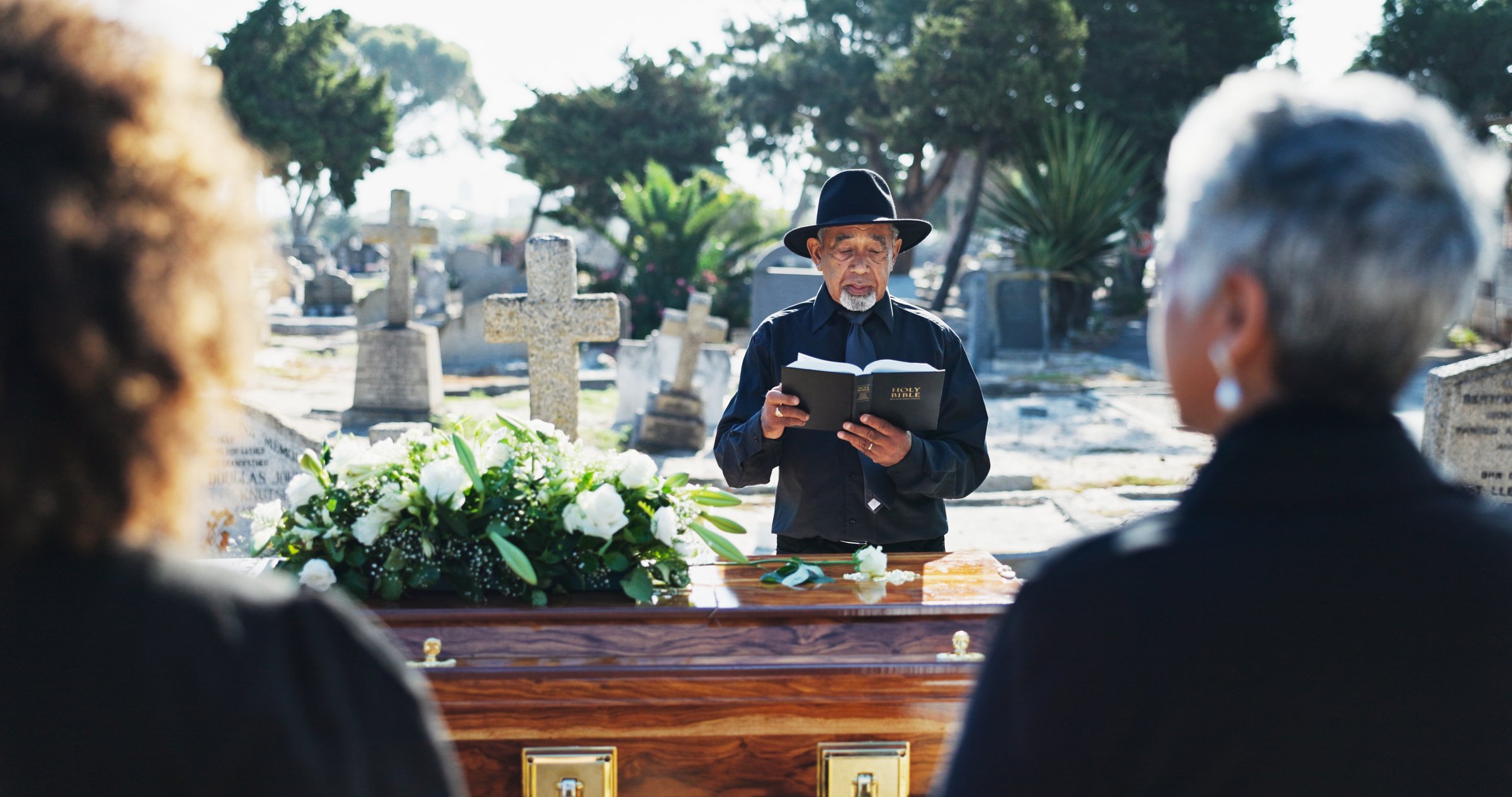 Bible, coffin and pastor by graveyard with family for spiritual farewell, grief or loss. Religion, casket and priest with people for memorial service at burial with holy book for blessing or goodbye.