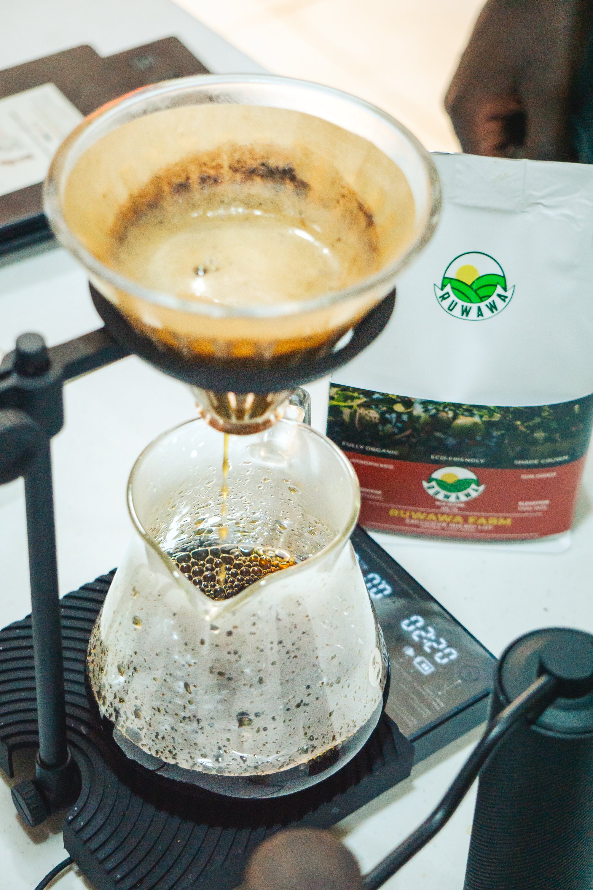 Pour-over coffee setup with brewed coffee dripping into a glass carafe, beside a bag of Suwawa coffee on a digital scale.