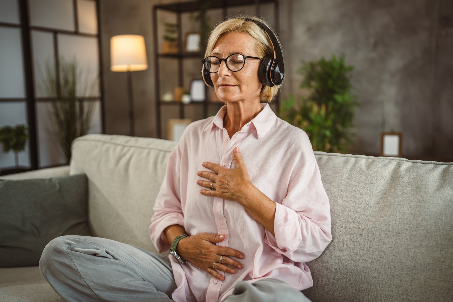 Mature woman sit on sofa with headphones and have guided meditation