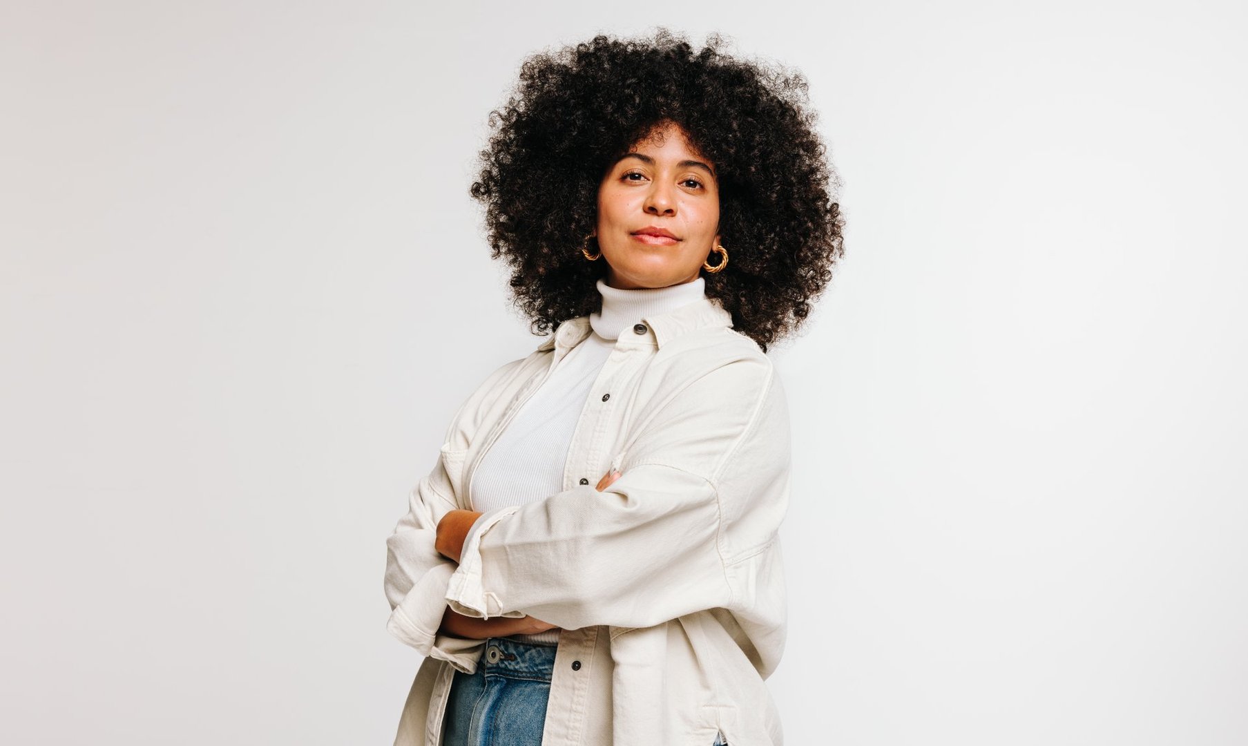 Self-confident woman with an Afro hairstyle looking at the camera while standing against a studio background. Attractive young woman of colour wearing her curly hair with pride.