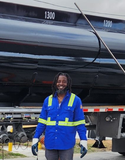 Man in a blue safety uniform with reflective stripes smiling in front of a parked tank trailer.