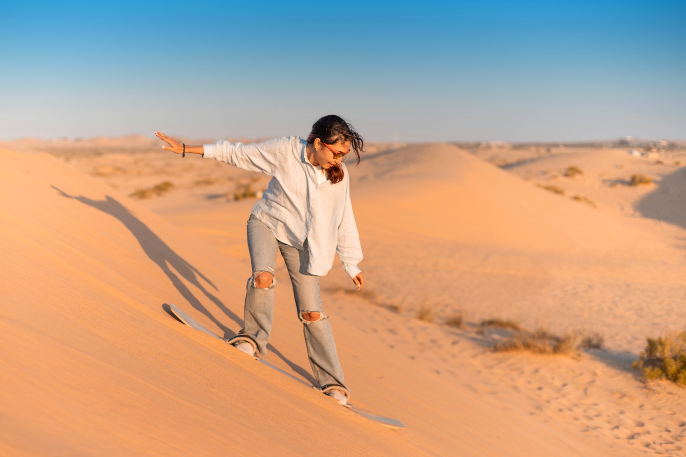 Young woman sandboarding down a sandy dune in the desert, enjoying the thrill of the sport during a stunning sunset