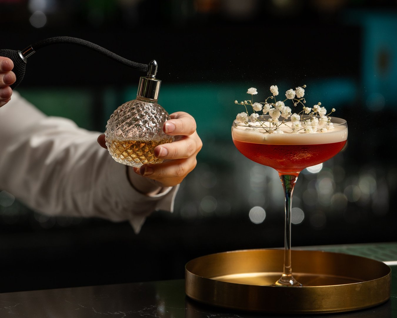A bartender's hand is delicately misting a blush-toned, floral-garnished cocktail with a vintage perfume atomizer, adding an aromatic layer to the drink's presentation