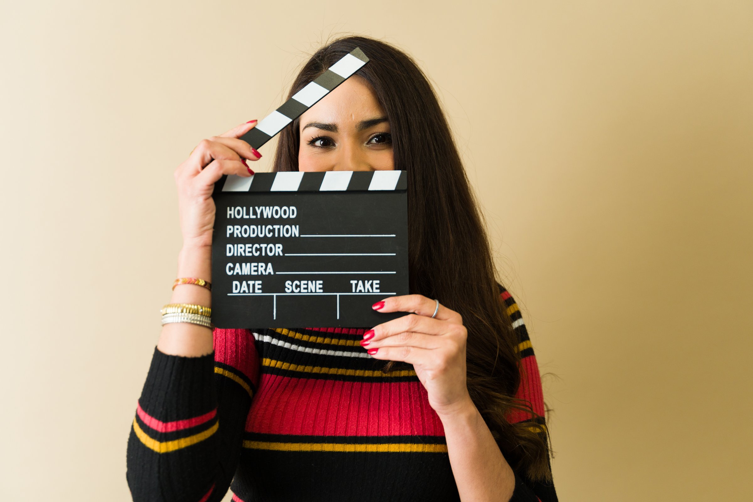 Young smiling latin filmmaker playfully holding a hollywood clapperboard in front of her face, radiating joy and enthusiasm for cinema