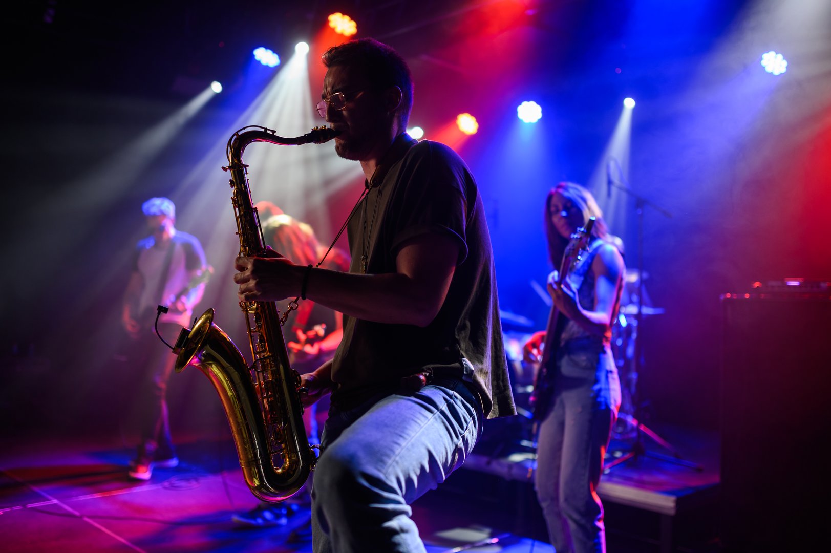 Saxophonist playing with energy on stage alongside band members under vibrant, colorful stage lights during a live musical performance