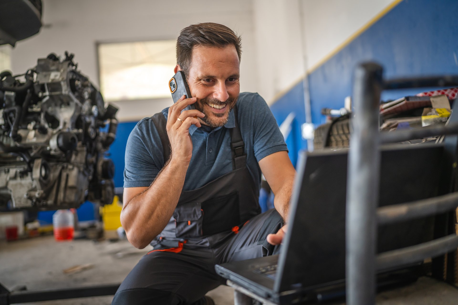 A car mechanic is multitasking, work on his laptop while engaged in a mobile phone call. He is in a garage setting, highlighting modern technology in auto repairs