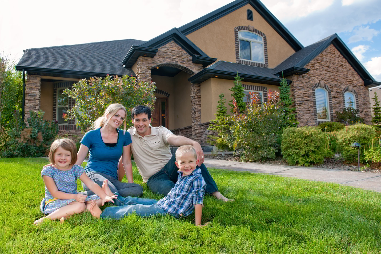 Family of four, including mother, father, and son and daughter sits in front of their beautiful home. They are proud and smiling to be in such a great place.