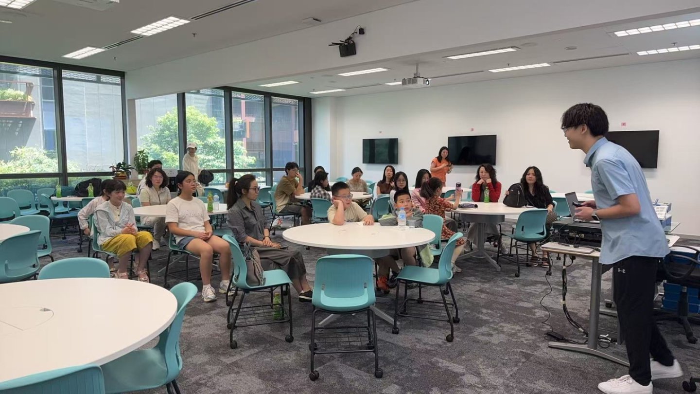 A group of people seated around tables in a classroom, listening to a person standing at the front, giving a presentation.