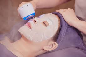 Person receiving a facial treatment with a sheet mask and a handheld blue light device at a spa.