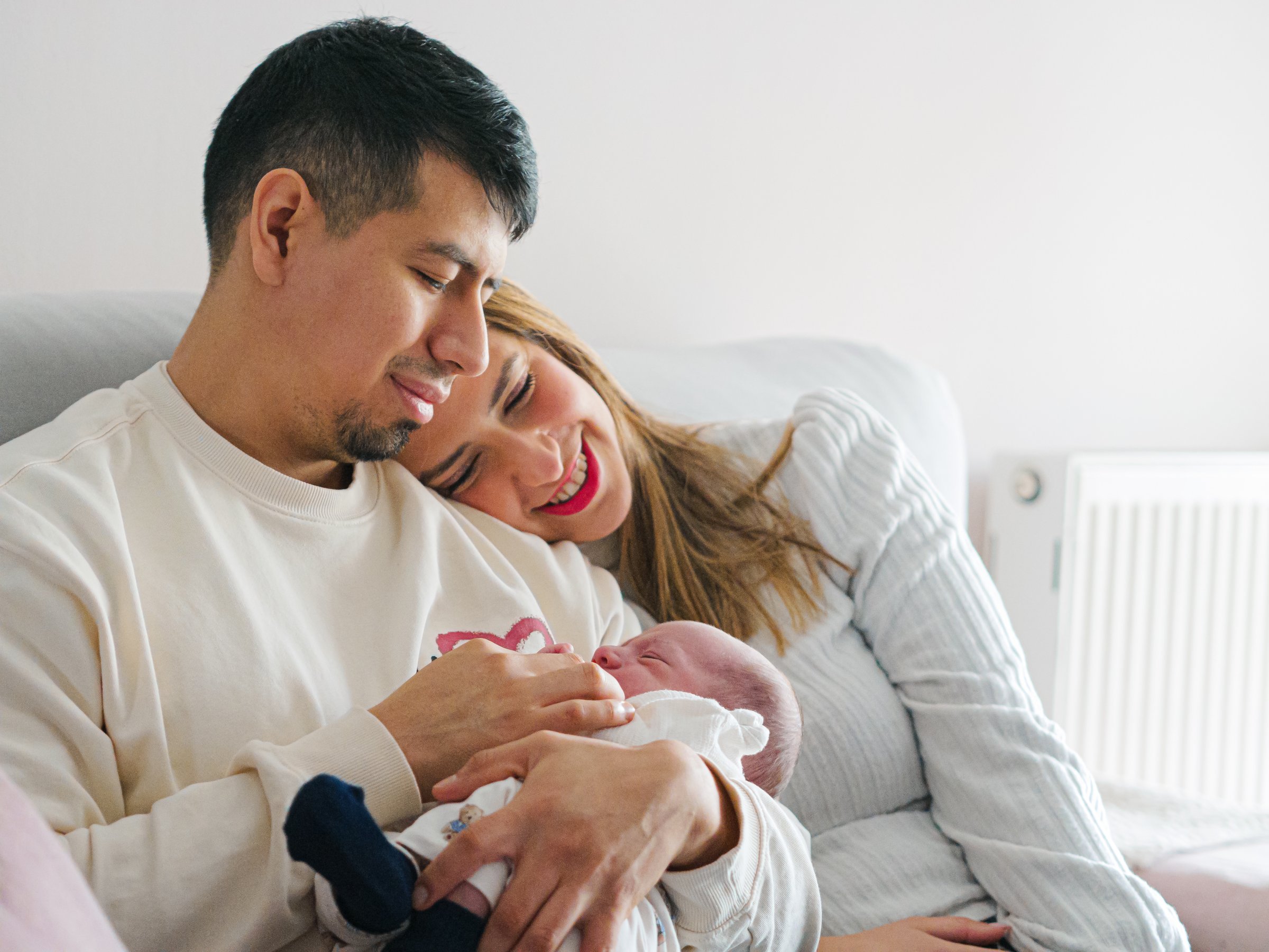Young adult parents look their baby sleep in arms whit love while sitting on the sofa of home. Portrait of diverse and multicultural family.