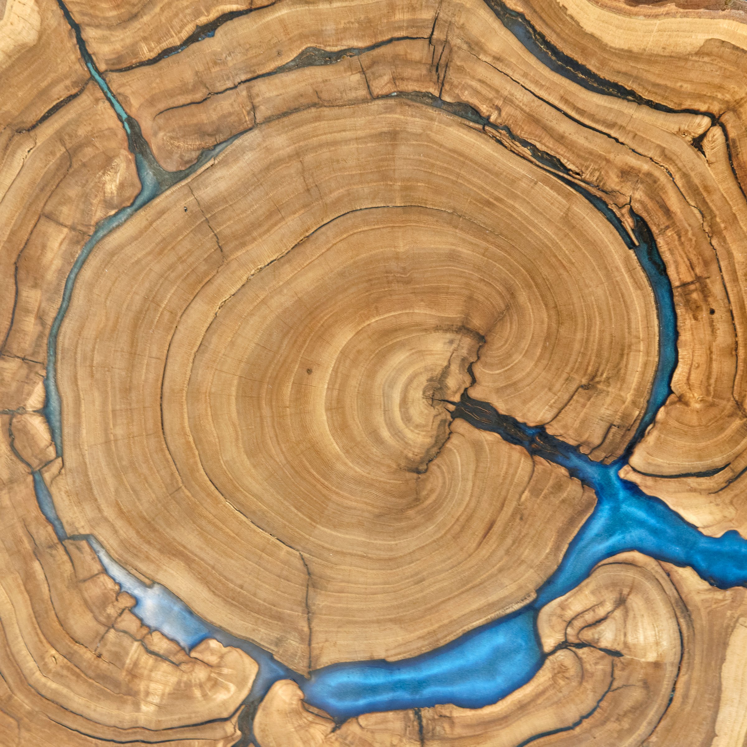 Close-up of wooden cross-section, tabletop, with natural rings and cracks filled with blue epoxy resin, creating striking contrast between organic and modern design elements, as abstract background