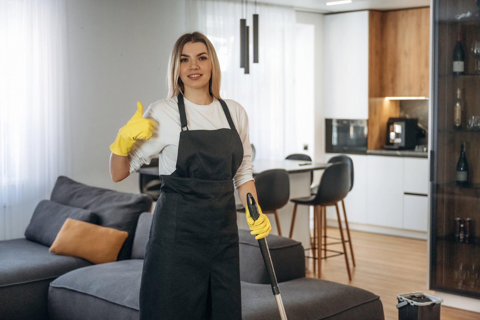 With thumb up, standing. Female professional cleaning service worker is domestic room.