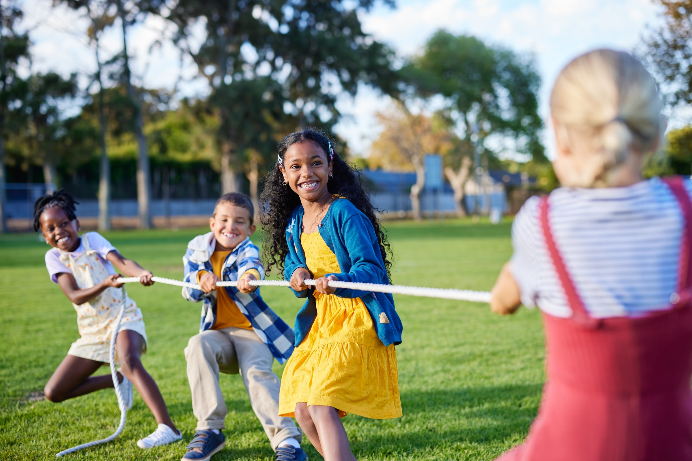 Group of happy multiethnic school kids playing tug of war with rope in city park. Little girls and boys playing tug of war in school park during break. Group of children playing together at summer camp.