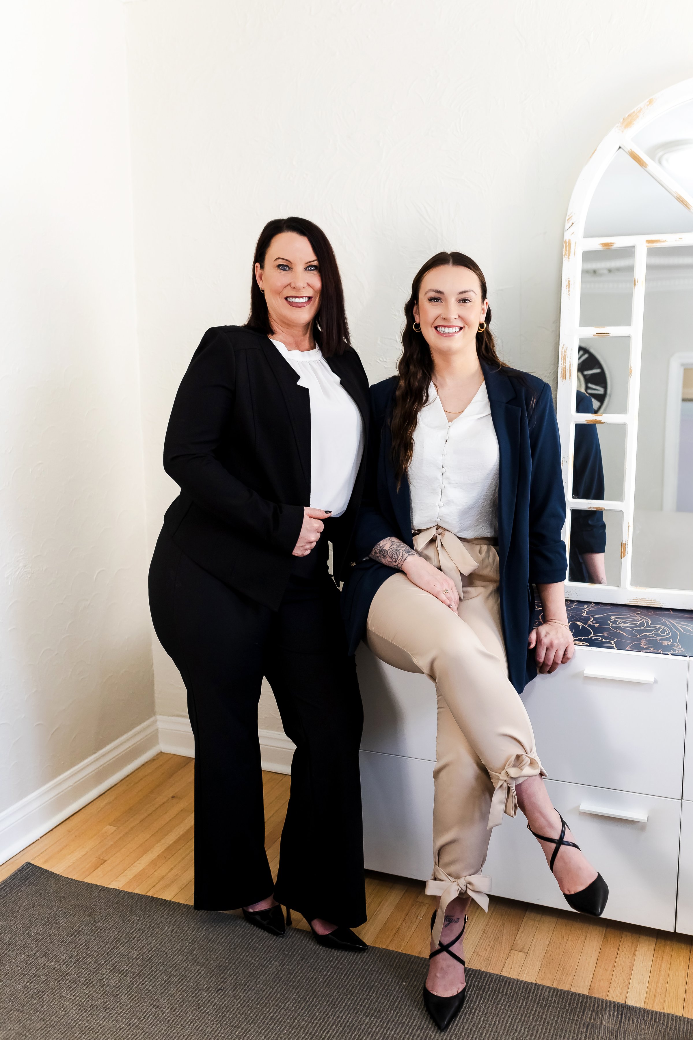 Two women in business attire, smiling, pose in a bright office setting with a mirror and white furniture.