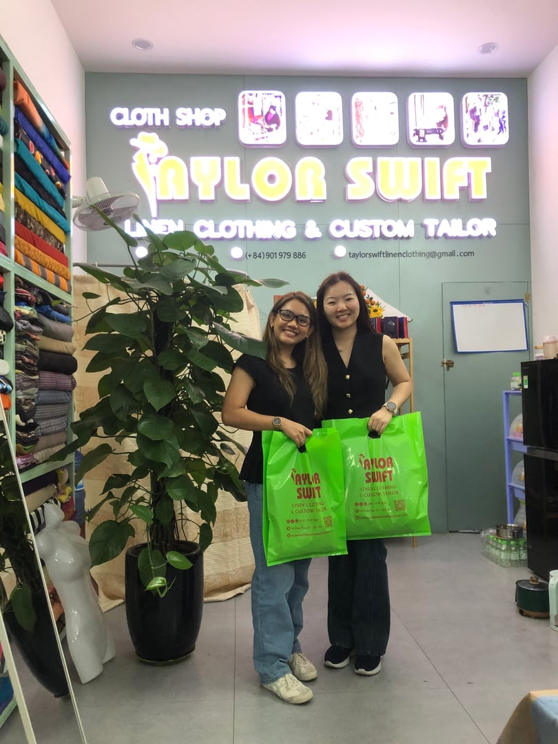 Two women smiling, holding green bags, inside "Taylor Swift" clothing shop with fabric rolls in the background.