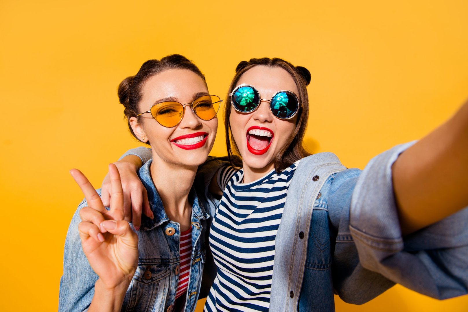Two glad positive grinning lady stand in glasses spectacles street style stylish trendy cool casual denim jeans clothes isolated on yellow background in take picture on cellular make hollywood smile