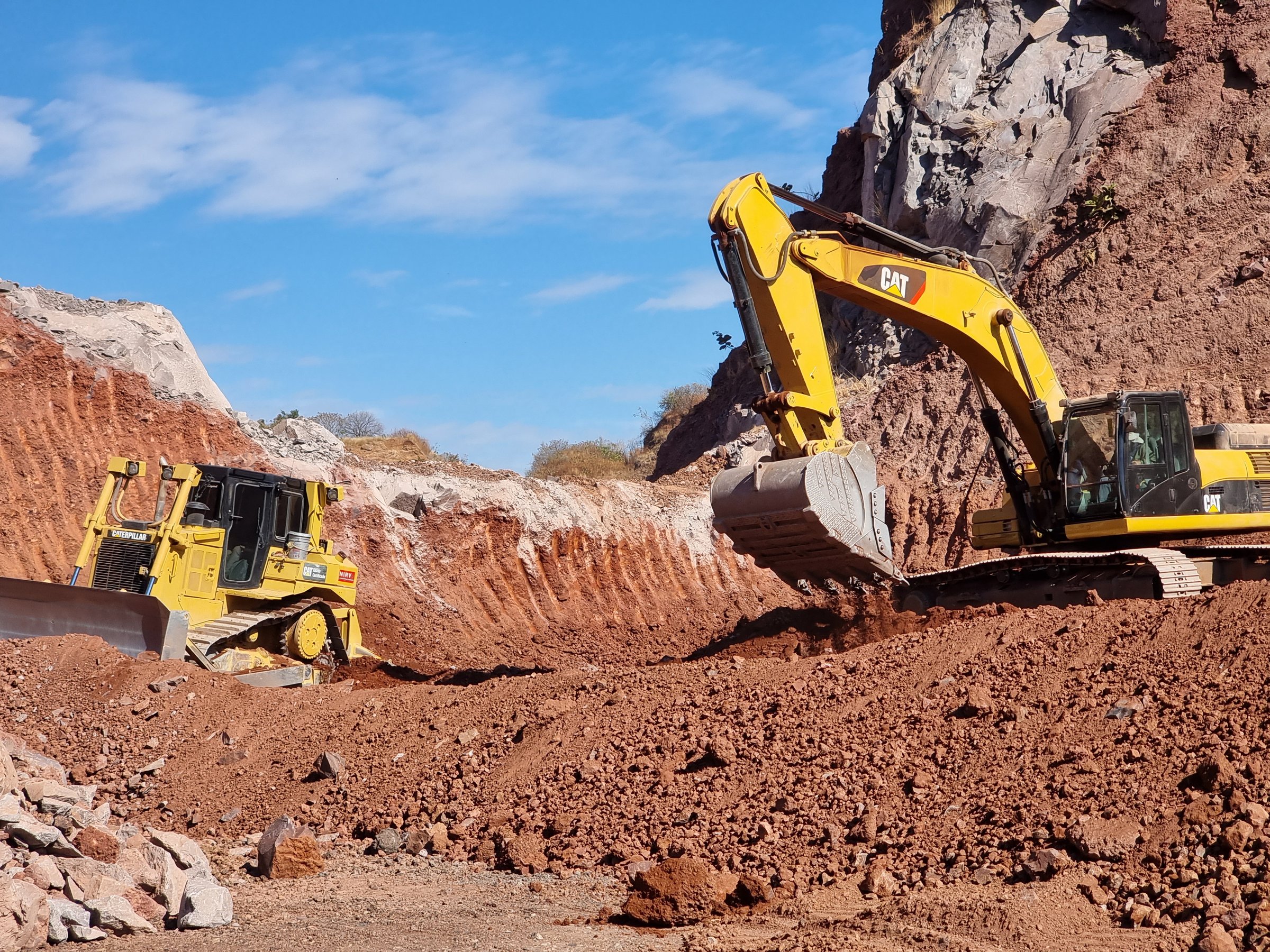 Excavators and bulldozers working on a construction site with red earth and rocky terrain under a clear blue sky.