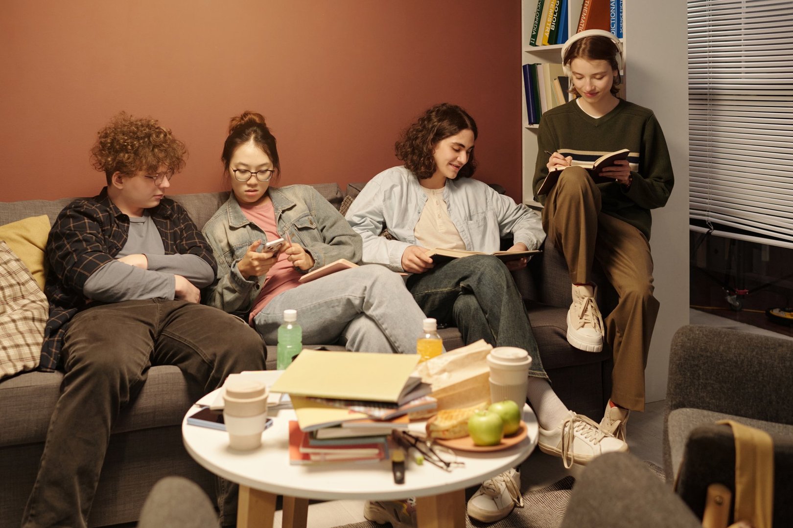 Group of teenage students relaxing together sitting on couch in cozy campus lounge doing homework and using gadgets in evening with low light