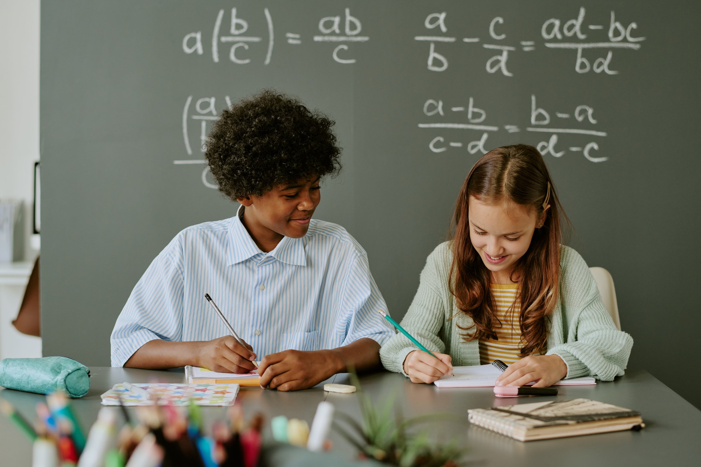 Caucasian girl and Black boy sitting at desk writing in notebooks, smiling and studying together in classroom with mathematical equations on chalkboard in background