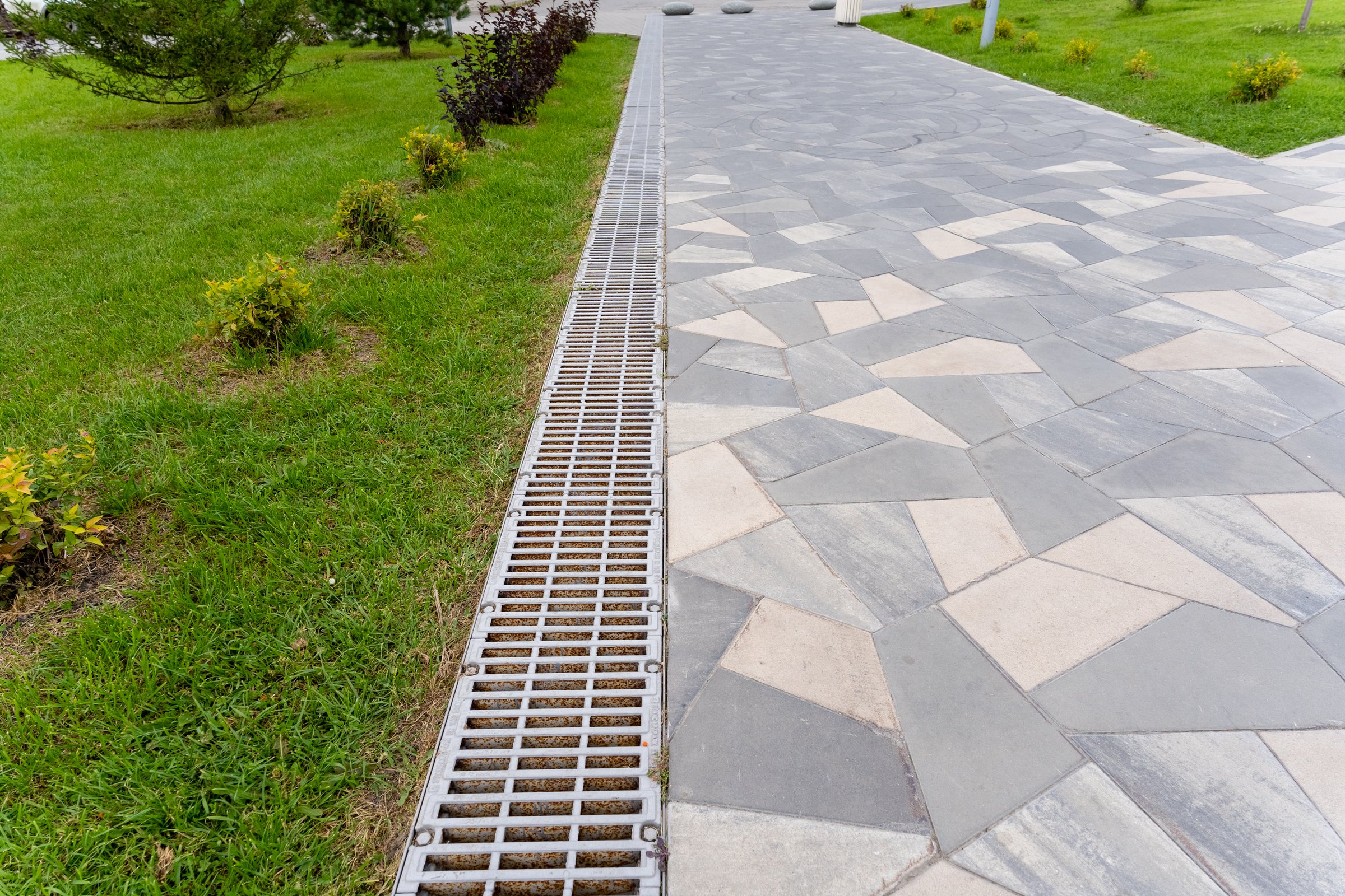 A metal drain is located strategically on the side of a brick walkway within a beautifully designed park, ensuring proper drainage